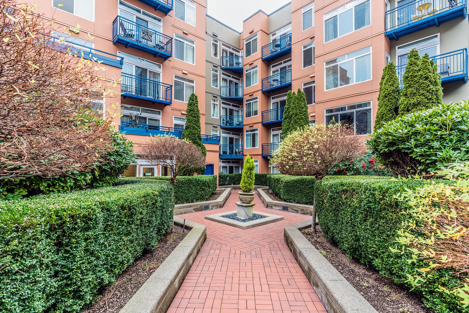 A multi-story residential building with balconies, a central pathway, and well-maintained hedges.