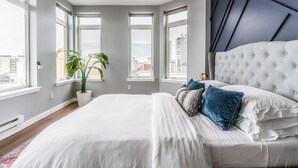 A modern bedroom with a large bed, a potted plant, and multiple windows offering city views. - Cozy Belltown Condo with amazing Elliott Bay Views (Seattle)