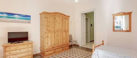 A bedroom with a wooden wardrobe, a television on a dresser, a bed with a white bedspread, and a patterned floor.