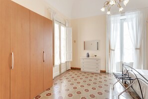 A bedroom with a wooden wardrobe, a white dresser, a bed with a metal frame, and a patterned floor.