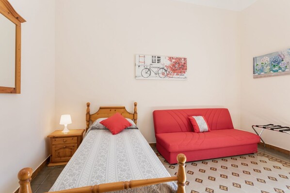 A bedroom with a bed, a red sofa, a nightstand with a lamp, and two wall art pieces.