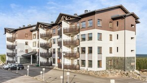 A multi-story residential building with balconies, a parking area, and a view of the landscape.