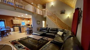 A spacious living area with a large black leather sofa, a wooden coffee table, and a patterned rug. A kitchen with wooden cabinets and a dining area with a table and chairs are visible in the background. - Riverfront Mtn Retreat Hot Tub/Fireplaces/King Beds/Tubing/Fishing/Spacious (Ellijay)