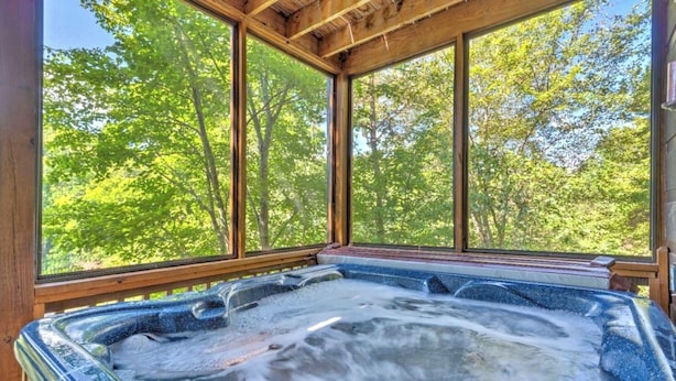 A hot tub with a wooden deck and large windows overlooking a forest. - Riverfront Mtn Retreat Hot Tub/Fireplaces/King Beds/Tubing/Fishing/Spacious (Ellijay)