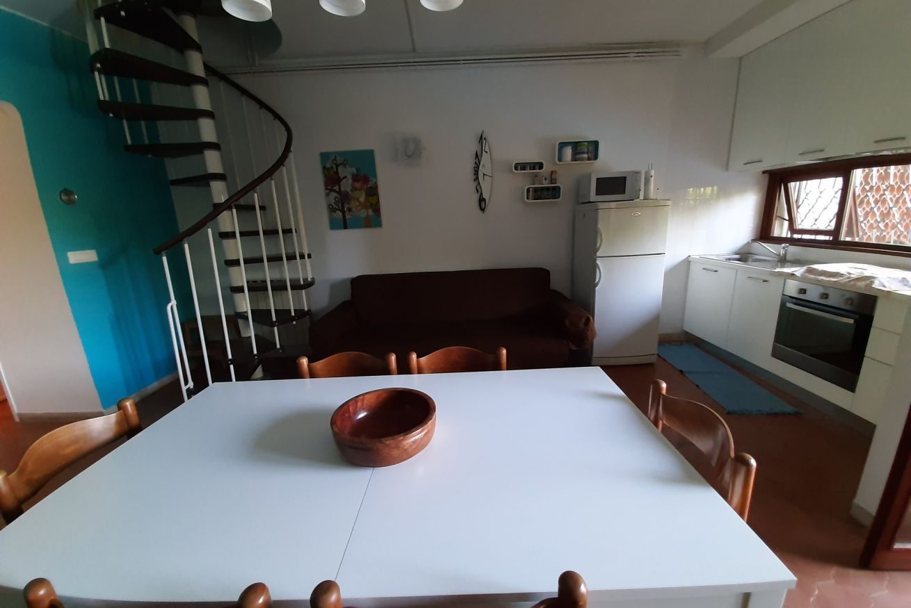 A dining area with a white table, brown chairs, a brown bowl, a spiral staircase, a kitchen with white cabinets, a microwave, and an oven.