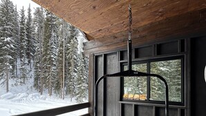 Una terraza de madera con una mecedora colgante, una pequeña mesa redonda y vistas a un bosque nevado a través de una ventana.