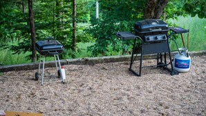 Een draagbare grill en propaantank staan op een grindpad, met bebossing op de achtergrond.