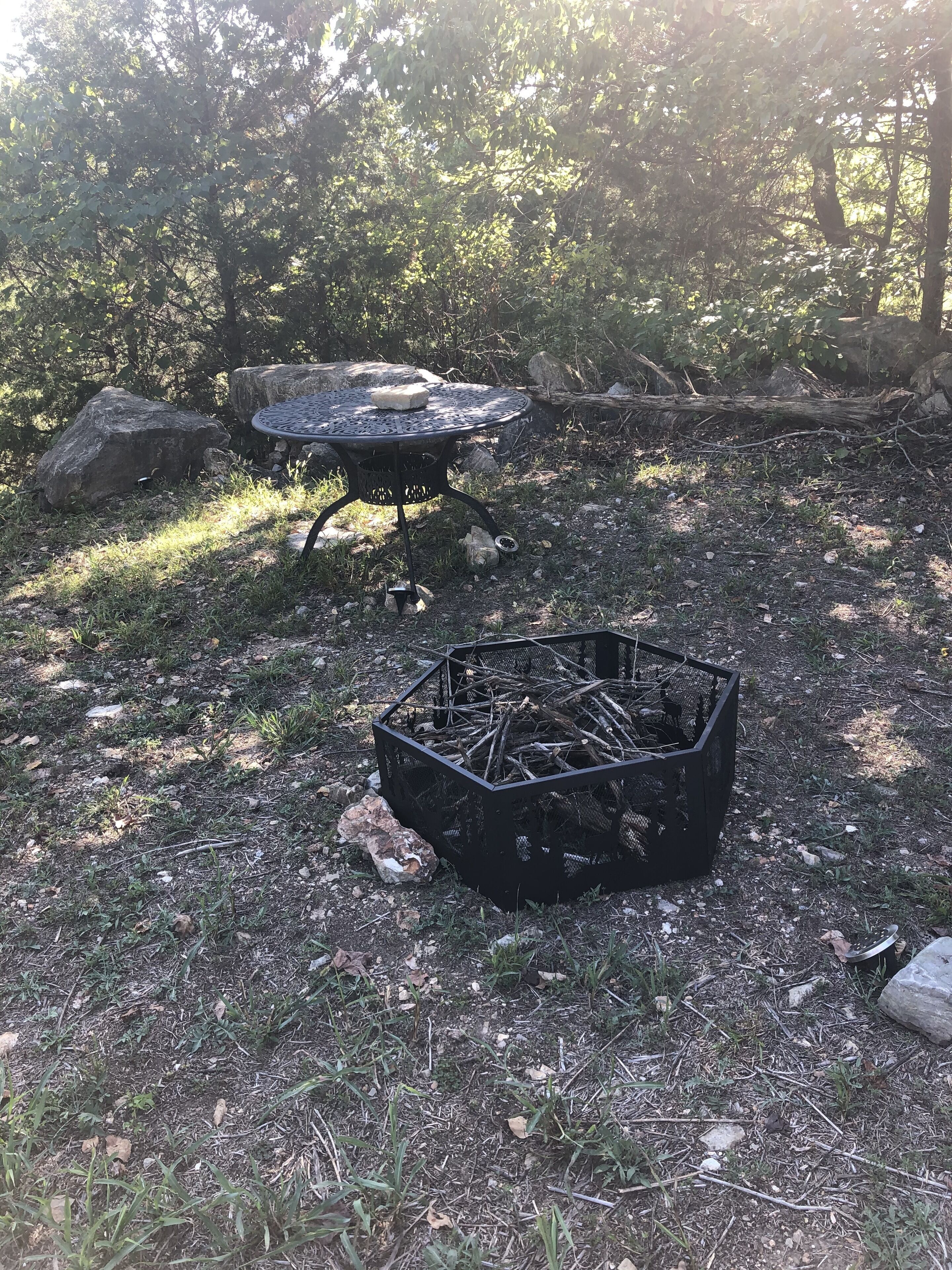 A metal table with a fire pit in the center, surrounded by rocks and trees.