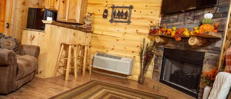 A cozy log cabin interior with a fireplace, wooden walls, a sofa, a ladder, and a TV mounted above the fireplace.