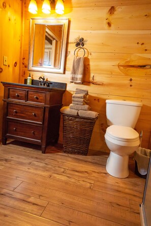 A wooden cabin bathroom with a wooden vanity, a white toilet, and a wicker basket with towels. - Hunting Cabin w/ Air Purifier (Branson)