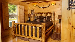 Un dormitorio de cabaña de madera con cama, dos lámparas de pared, decoración rústica y ventana con vistas.