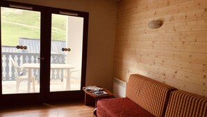Una habitación con piso de madera, una cama con colcha roja, una mesita con revistas y vistas a un área exterior con una mesa y sillas a través de una puerta de vidrio.