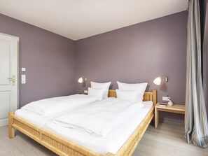 A double bed with white linens in a room with purple walls and a wooden headboard. There are two bedside tables with lamps, a door, and a striped curtain.