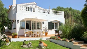 Una casa de dos pisos con un balcón, un patio con mobiliario exterior y un jardín con plantas en macetas.