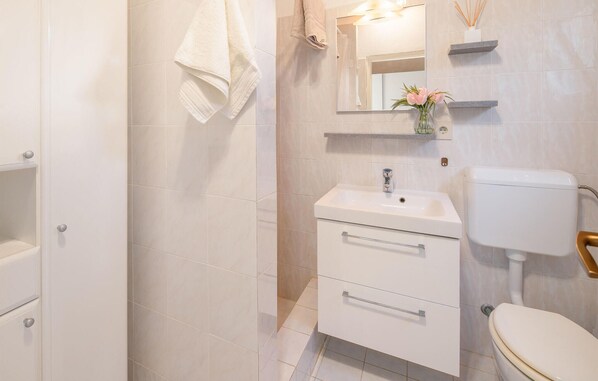 Une salle de bain moderne avec un lavabo, des toilettes et un meuble-lavabo blancs. Deux serviettes sont accrochées au mur, un miroir se trouve au-dessus du lavabo et une petite étagère supporte un vase de fleurs.