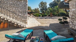 A stone building with a staircase, two lounge chairs with cushions, a table with a book and a teapot, and a garden with various plants and trees. - 1 bedroom amazing apartment in Ravni (Ravni)