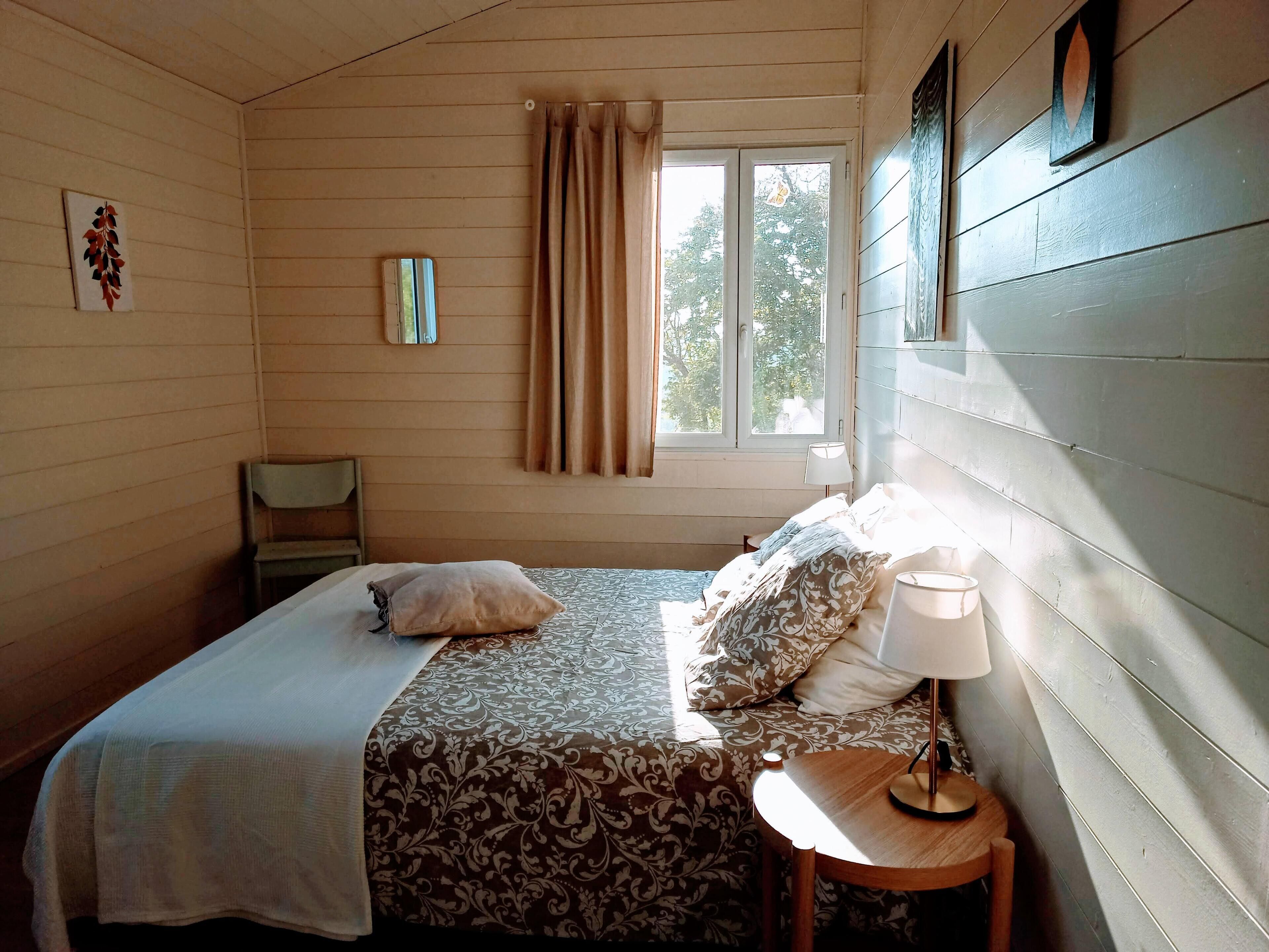 Ein Zimmer mit einem Bett, einem Nachttisch mit Lampe, einem Sessel, einem Fenster mit Vorhängen und einem Wandbild.
