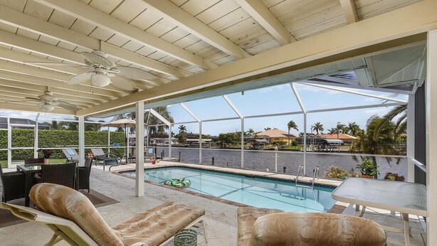 An outdoor pool area with lounge chairs and a table, overlooking a body of water and palm trees. - Holidays in Paradise! NO bridges, NO locks to the Gulf of Mexico! (Cape Coral)