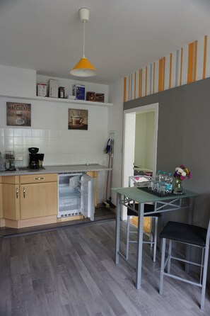 Eine kleine Küche mit weißem Arbeitsplatz, einer Kaffeemaschine und einem Kühlschrank. Ein kleiner Tisch mit zwei Hockern und eine Blumenvase stehen dort.