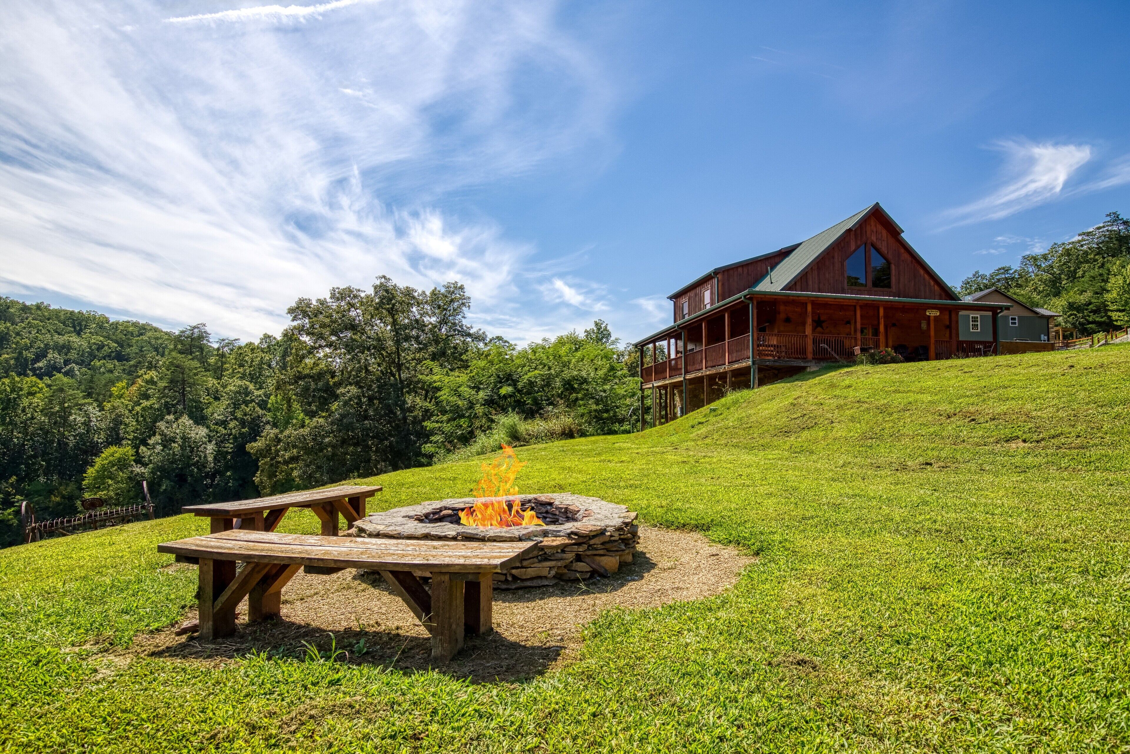 Eine rustikale Lodge mit Steinkamin und Picknicktischen auf einer grasbewachsenen Anhöhe, umgeben von Bäumen und blauem Himmel.