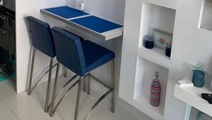 Una cocina compacta con encimera blanca, dos sillas azules y un cuadro abstracto azul en la pared.