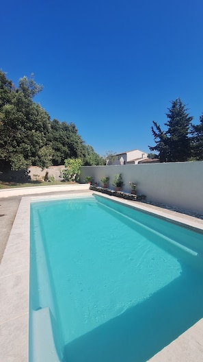 A rectangular swimming pool with clear blue water, surrounded by a low stone wall and a tall hedge.