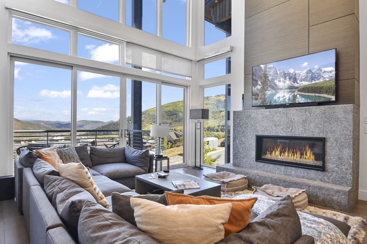 A modern living room with a large sectional sofa, a fireplace, a flat-screen TV, and a view of mountains and a lake through large windows.