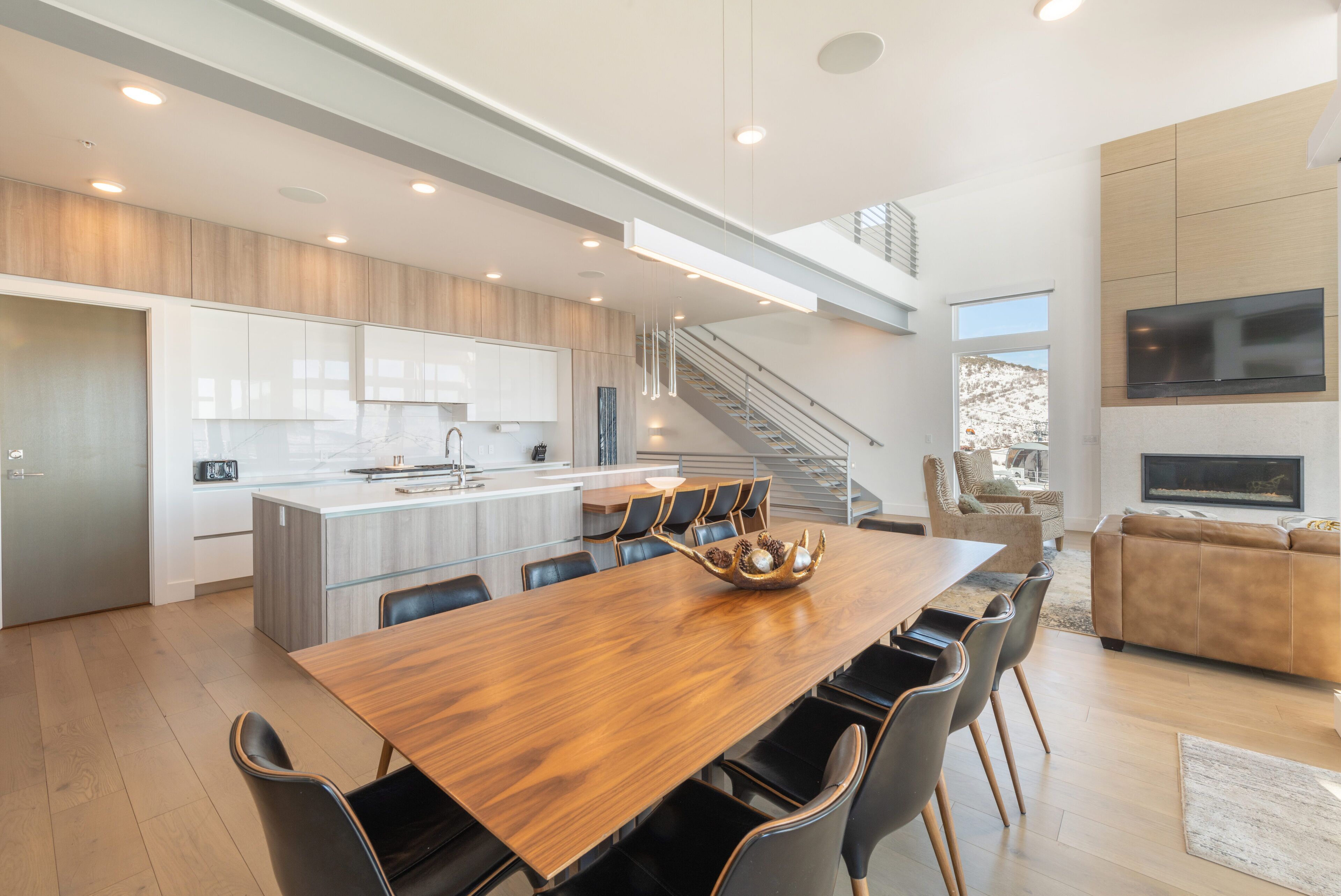 A spacious modern kitchen with a large island, a dining area with a wooden table and chairs, a staircase leading to an upper level, and a living area with a sofa and TV.