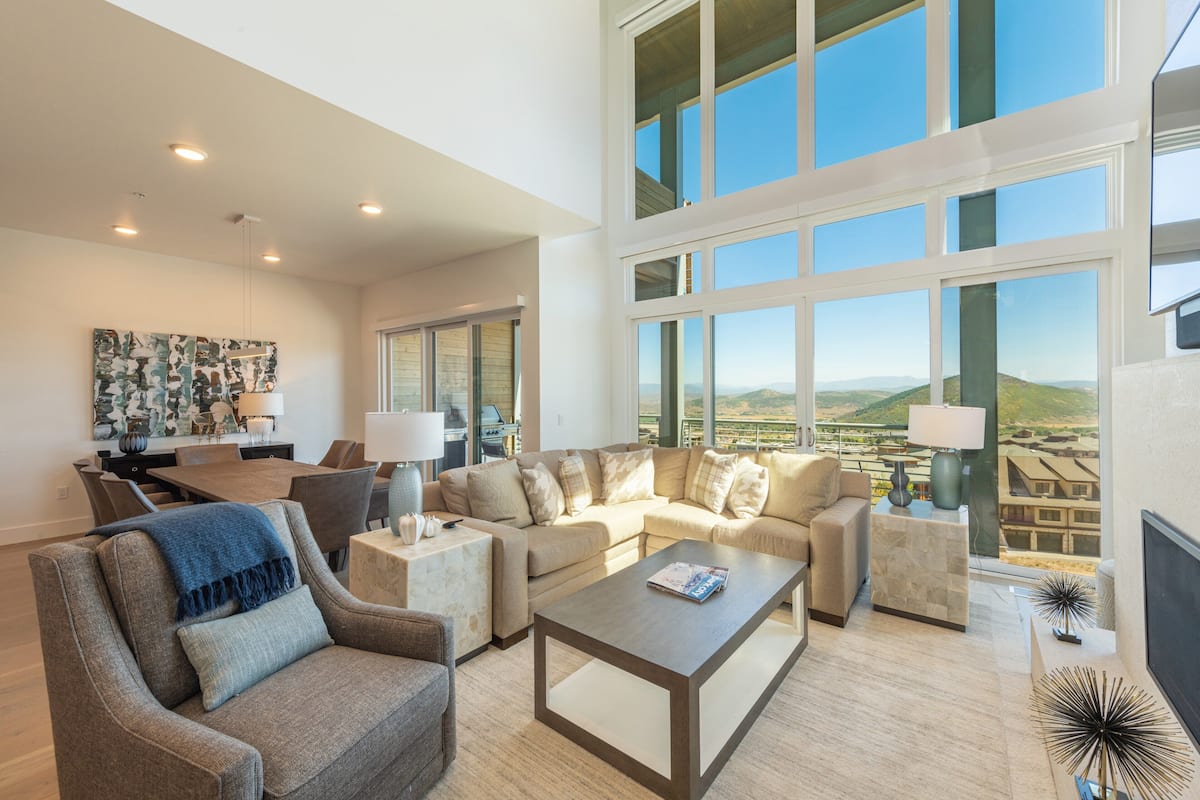A spacious living room with a large window offering a view of the countryside. There is a dining area with a table and chairs, and a sofa set.