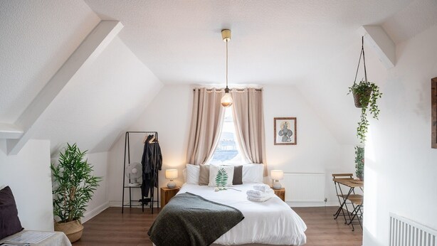 Une chambre à coucher avec un lit, des lampes de chevet, un radiateur, une plante suspendue, un porte-manteau et une fenêtre avec des rideaux.