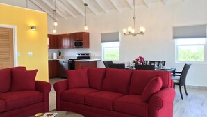 A modern living room with red sofas, a glass coffee table, and a kitchen in the background. - Tranquil Villa near Long Bay Beach, Pool & Sunsets (Long Bay Hills)