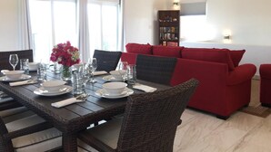 A dining area with a dark wooden table set for four, surrounded by wicker chairs. A red sofa and a bookshelf with books are in the background. - Tranquil Villa near Long Bay Beach, Pool & Sunsets (Long Bay Hills)