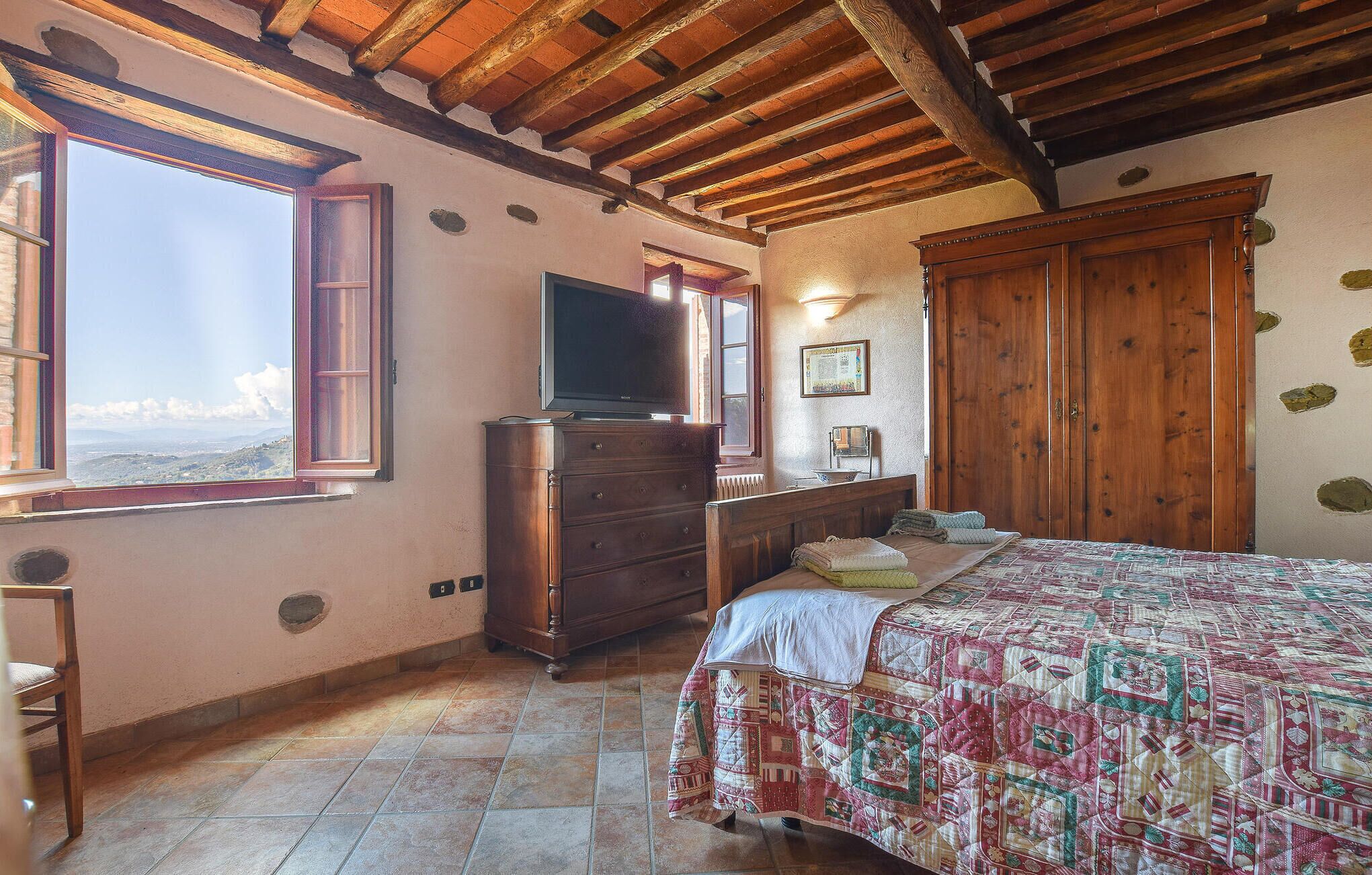 A bedroom with a wooden ceiling, a large window with a view, a bed with a quilt, a dresser, and a television.