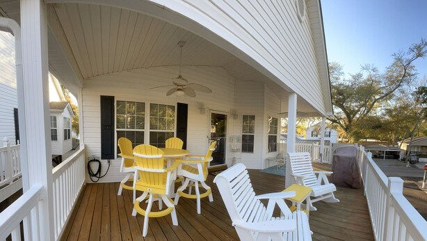 Une véranda couverte avec une terrasse en bois, des rampes blanches et du mobilier d’extérieur comprenant des chaises et un fauteuil à bascule.