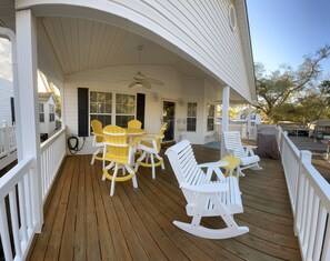 Une véranda couverte avec une terrasse en bois, des rampes blanches et du mobilier d’extérieur comprenant des chaises et un fauteuil à bascule.
