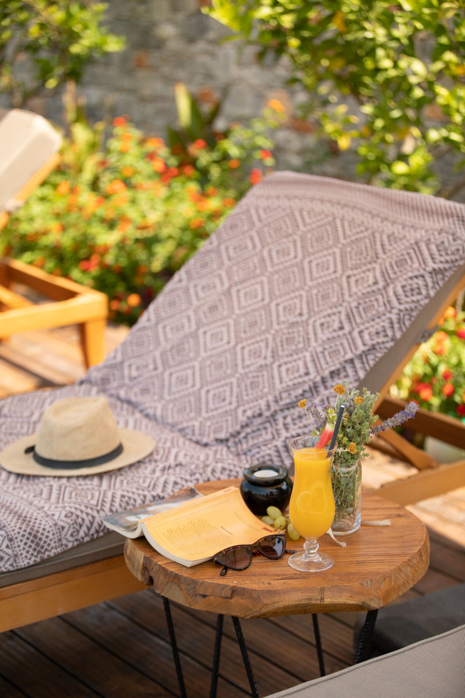 A sun lounger with a patterned cushion, a hat, a book, a pair of sunglasses, a glass of orange juice, and a small vase with flowers.