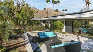 A modern outdoor living area with a sofa and chairs, surrounded by plants and palm trees, with a mountain in the background.