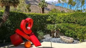 A landscaped area with a large red sculpture, palm trees, and a mountain in the background.