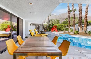 A modern outdoor pool area with a wooden table and yellow chairs, surrounded by palm trees and a mountain view.