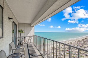A balcony with a view of the beach, chairs, and a small table.