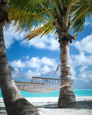A hammock tied between two palm trees on a sandy beach with clear blue water and a partly cloudy sky in the background.