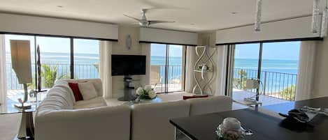 A modern living room with a sofa, a TV, and a balcony with a view of the ocean.