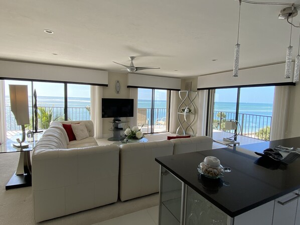 A modern living room with a sofa, a TV, and a balcony with a view of the ocean.