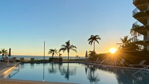 Ein Swimmingpool mit Liegestühlen und Palmen, mit Blick auf ein Gewässer bei Sonnenuntergang.