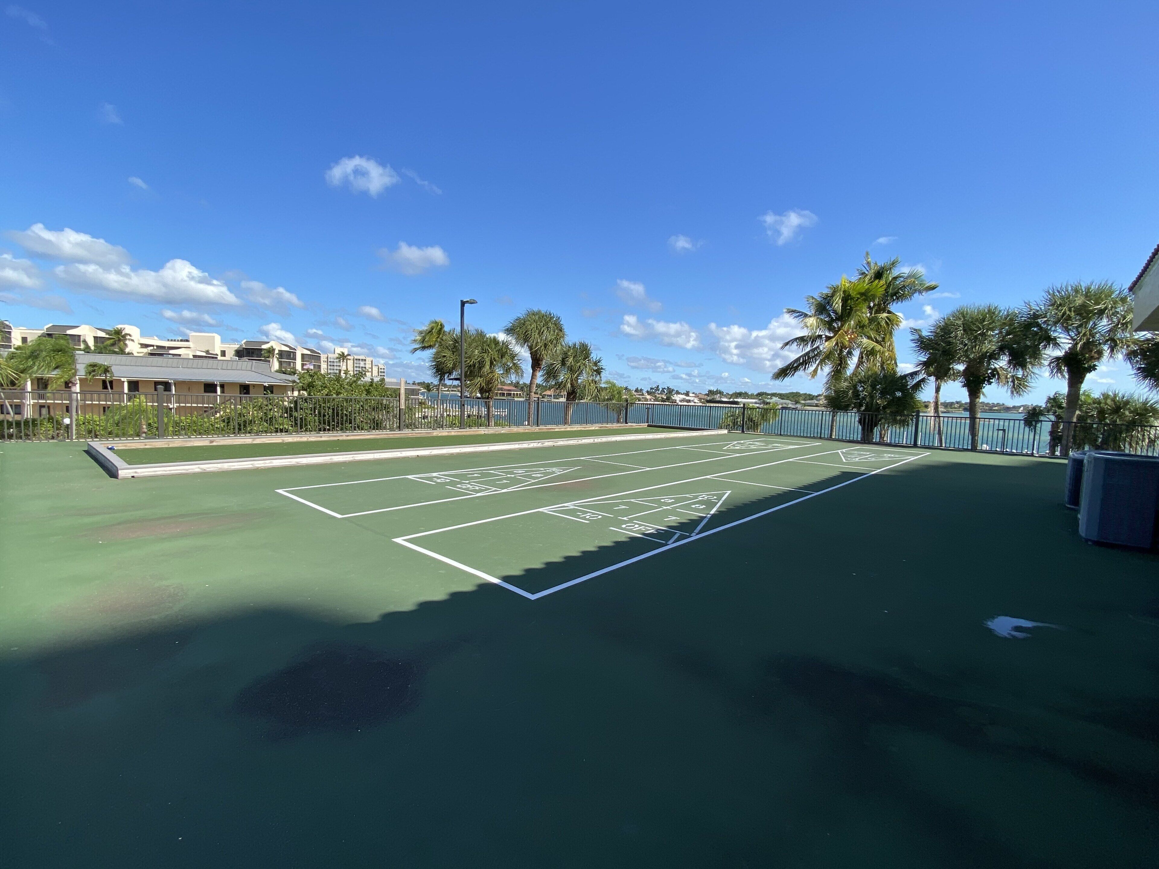 An outdoor tennis court with a green surface, surrounded by a fence and palm trees.