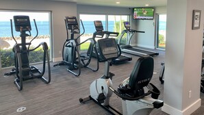 Ein Fitnessraum mit Laufbändern, Crosstrainern und einem Fahrradergometer mit Blick auf einen Strand.
