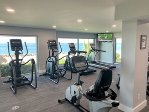 A gym with treadmills, elliptical machines, and a stationary bike, overlooking a beach.