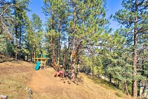 A playground with a slide and climbing structures, surrounded by tall pine trees.