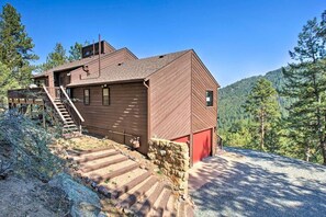 Uma cabana rústica de madeira com porta vermelha, escada que leva a um deck e vista para um morro arborizado.