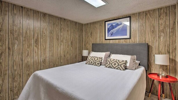 A hotel room with a wooden paneled wall, a bed with patterned pillows, a red side table, and a framed picture on the wall. - The Evergreen Gem| Mountain Experience Retreat (Evergreen)